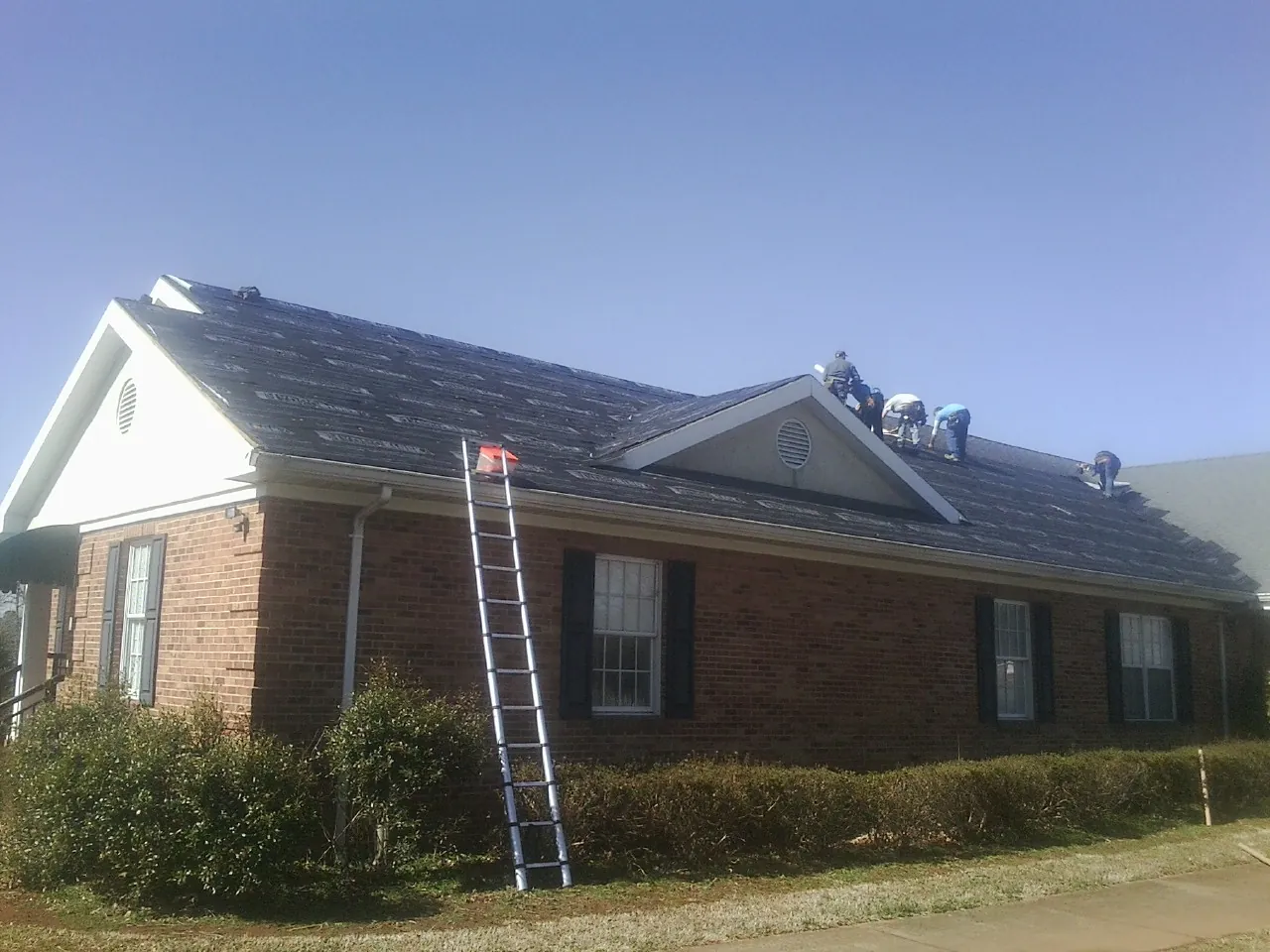Crew tearing shingles off a brick ranch roof with a ladder set — Emergency Roof Repair work by our crew in Mc Callsburg