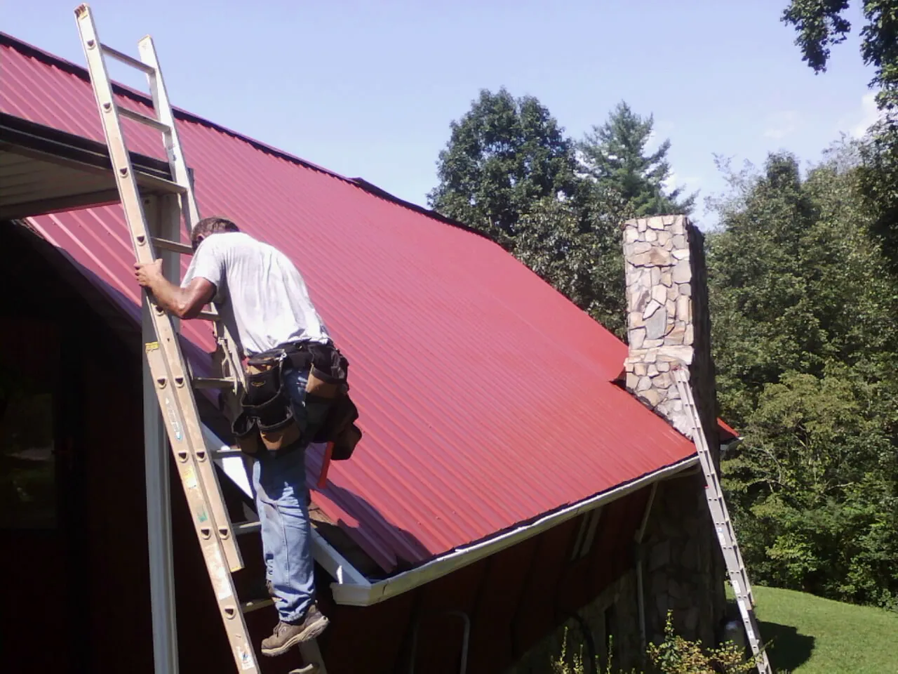 Roofer on a ladder beside a red metal roof and stone chimney — Soffit & Fascia Repair by Roofing Brothers in Mc Callsburg